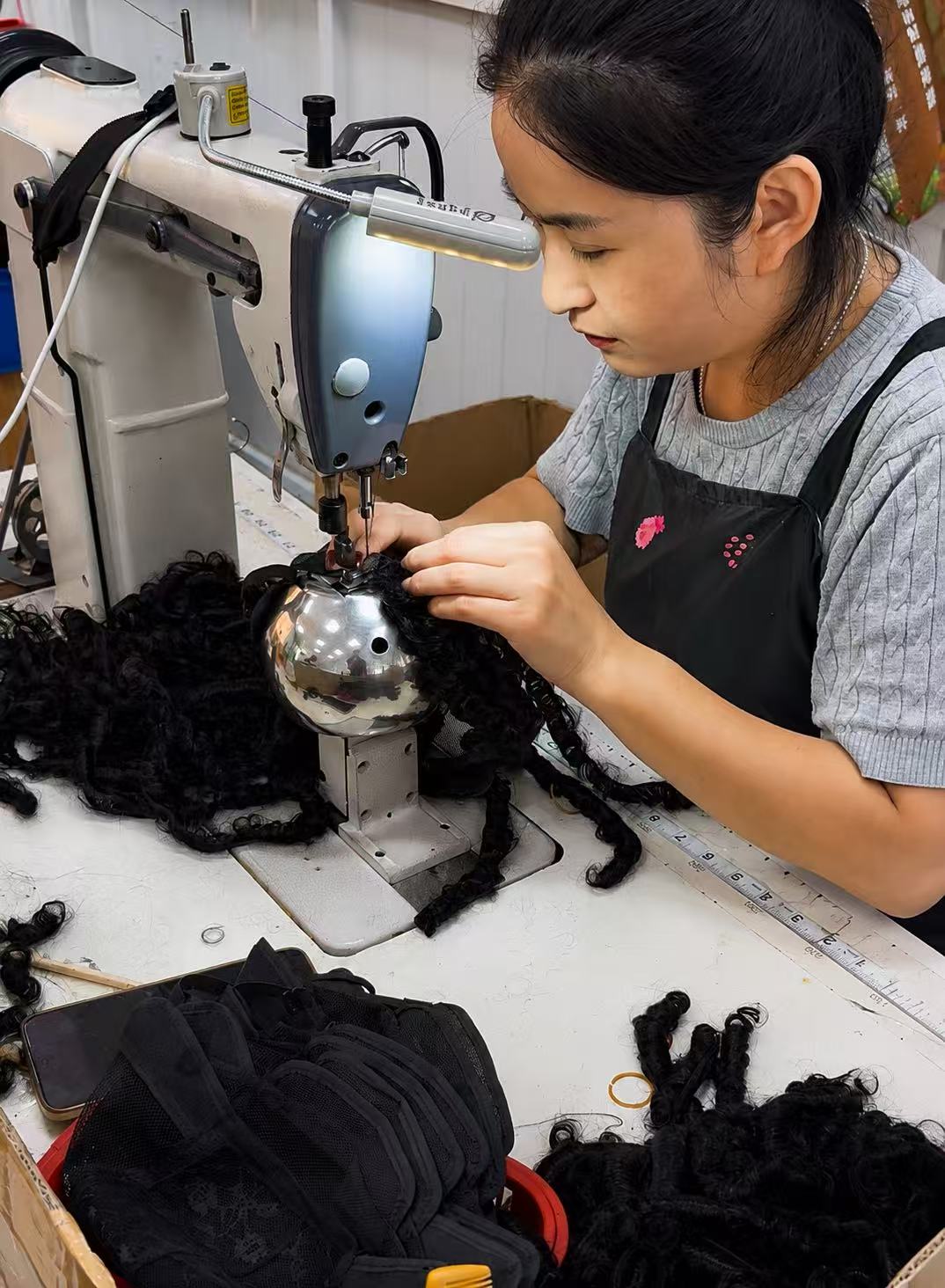 Production worker assembling curly hair and wig-cap components with an industrial sewing machine.