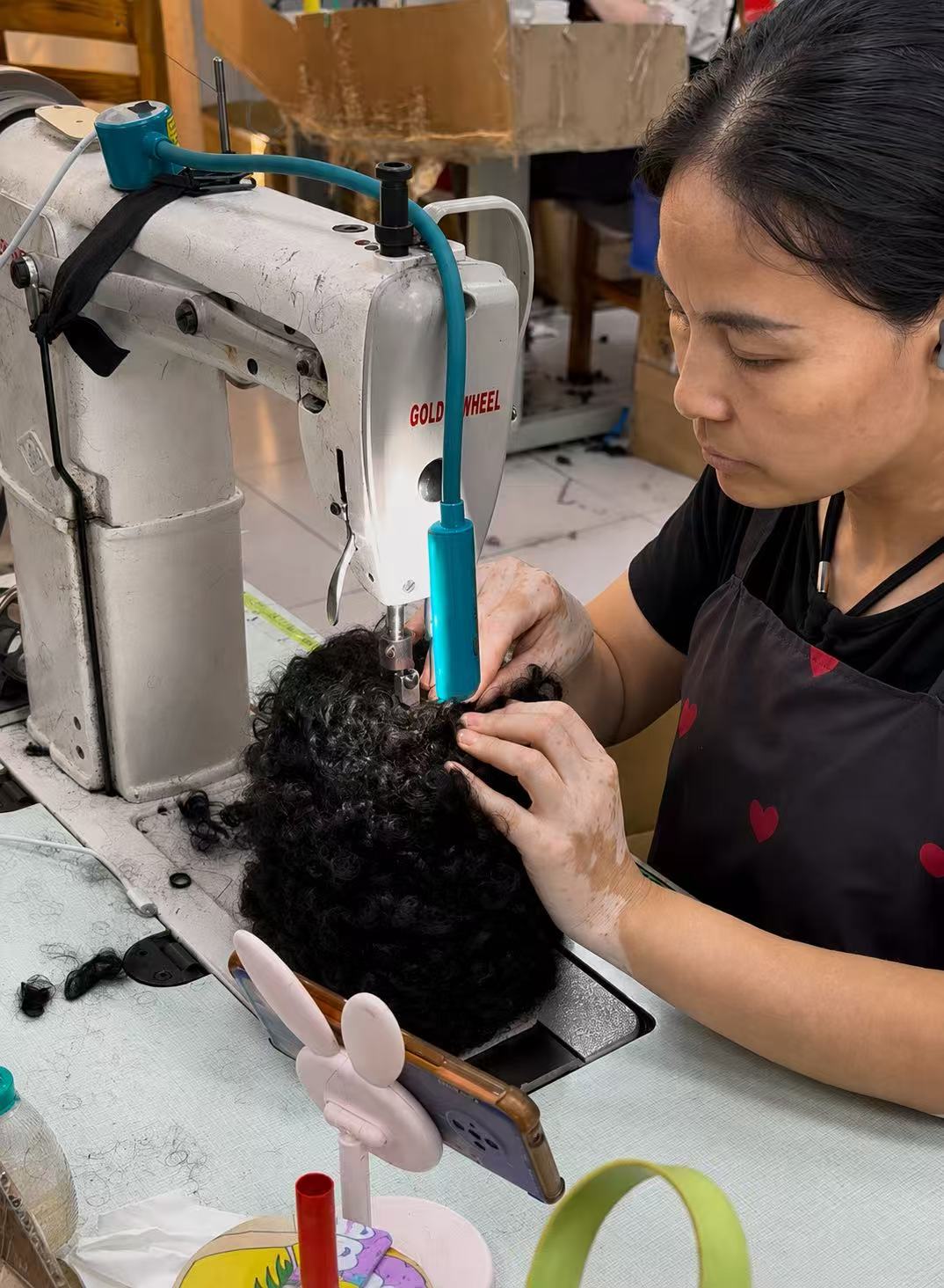 Production worker sewing curly hair onto a wig cap with an industrial machine.